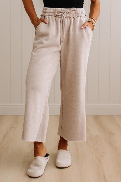 A person wearing light beige wide-leg cropped pants with an elastic drawstring waist, standing on a light wood floor against a white paneled wall.