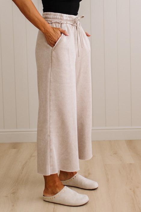 A person wearing light beige wide-leg cropped pants with a drawstring waist and side pockets, paired with white slip-on shoes, standing on a light wood floor against a white paneled wall.