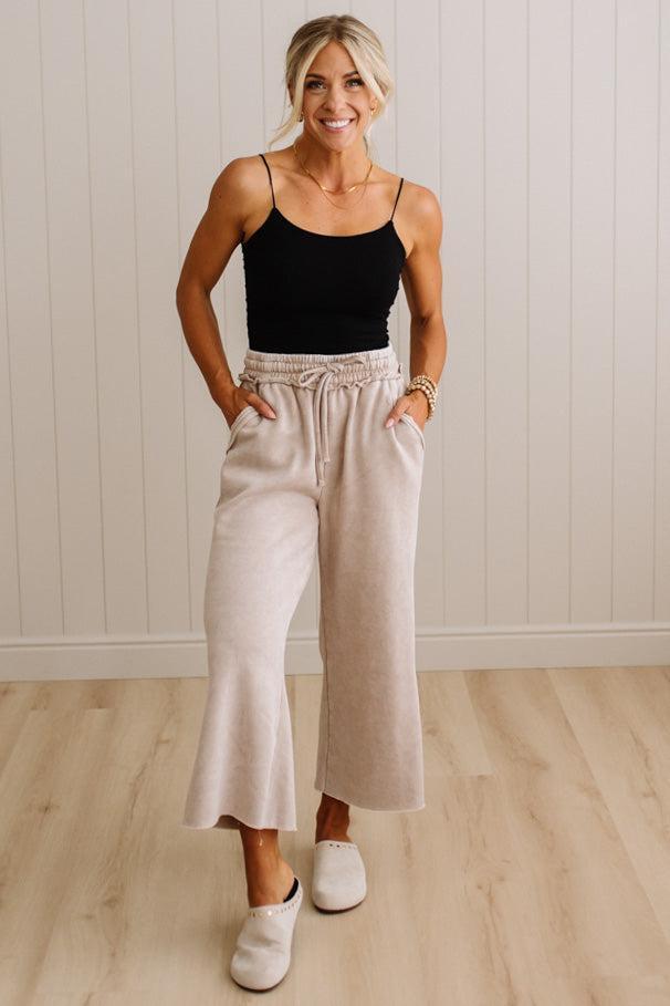 A woman modeling a pair of light beige wide-leg cropped pants with a drawstring waist, paired with a black camisole and white slip-on shoes, standing against a white paneled wall and light wood floor.