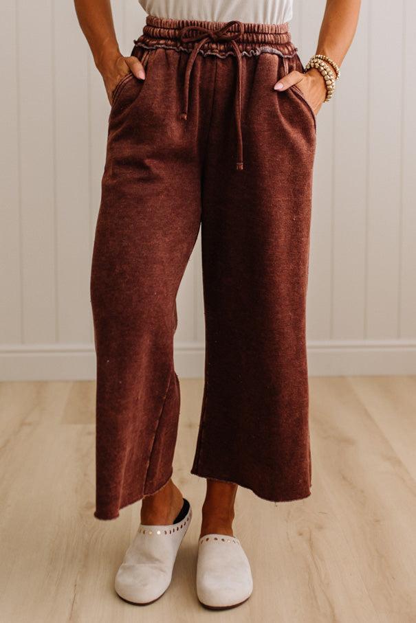 A person wearing rust-brown wide-leg cropped pants with an elastic drawstring waist, paired with white slip-on shoes, standing against a white paneled wall and light wood floor.