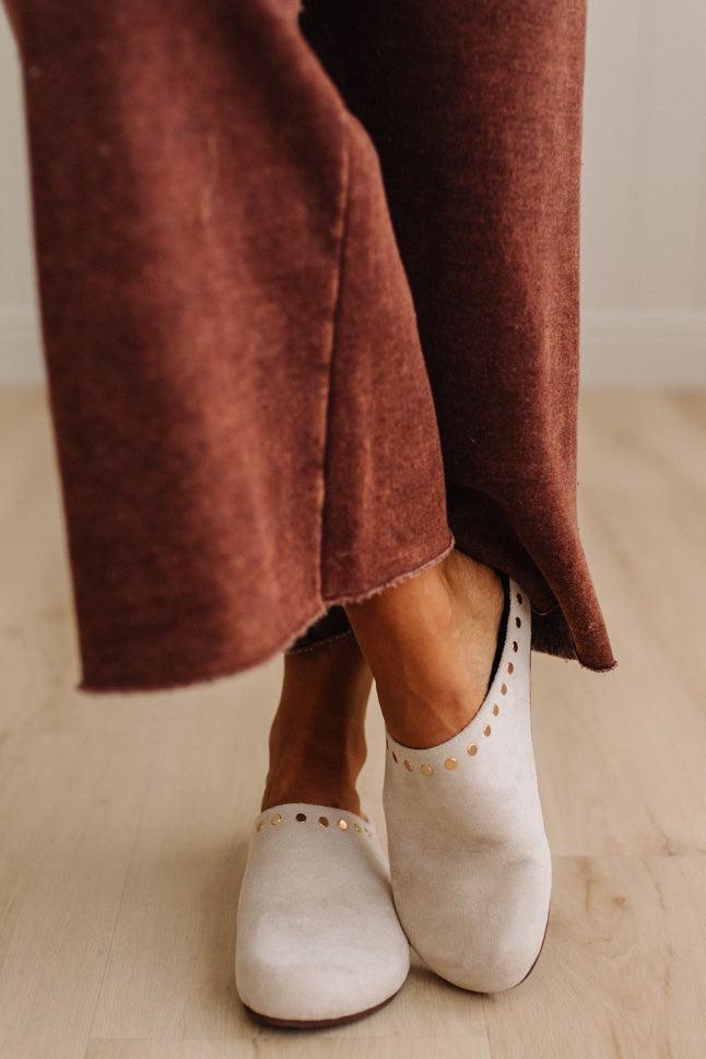 A close-up of a person's feet wearing off-white slip-on shoes with gold-tone stud detailing, paired with rust-brown wide-leg pants, standing on a light wood floor.