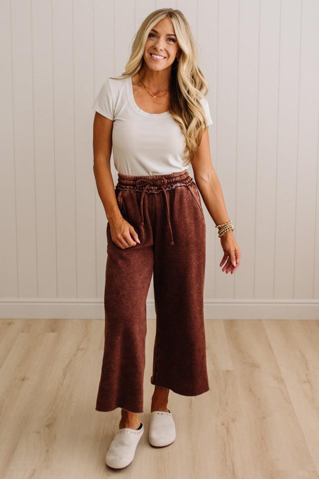 A woman modeling a casual outfit consisting of a white short-sleeve scoop-neck t-shirt and high-waisted, wide-leg brown pants with a drawstring waist, paired with white slip-on shoes, set against a light wood-paneled wall and light wood floor.