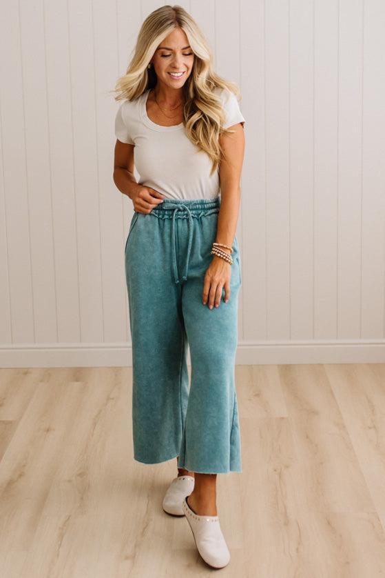 A woman modeling a pair of wide-leg cropped pants in a dusty teal color, paired with a white t-shirt and white slip-on shoes, standing in a room with light wood floors and white paneled walls.