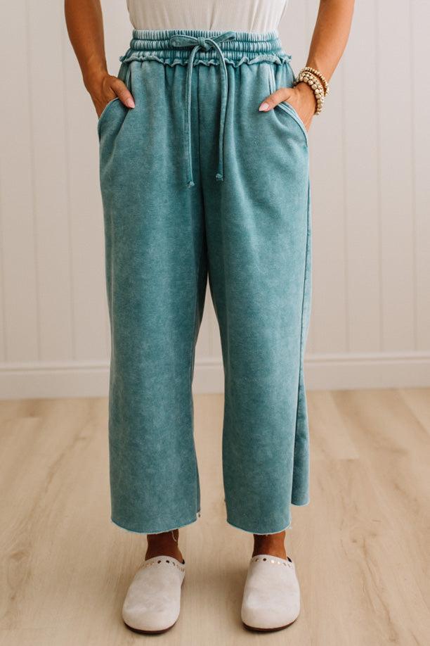 A person wearing wide-leg cropped pants in a heathered teal color, paired with a white top and white slip-on shoes, standing in a room with light wood floors and white paneled walls.