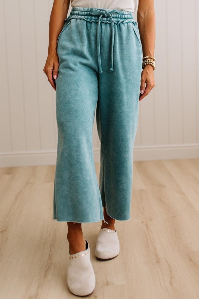 A person wearing high-waisted, wide-leg cropped pants in a dusty teal color, paired with white slip-on shoes, standing on a light wood floor against a white paneled wall.