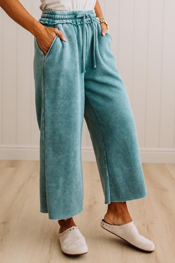 A person wearing wide-leg cropped pants in a dusty teal color, styled with white slip-on shoes against a light wood-paneled wall.