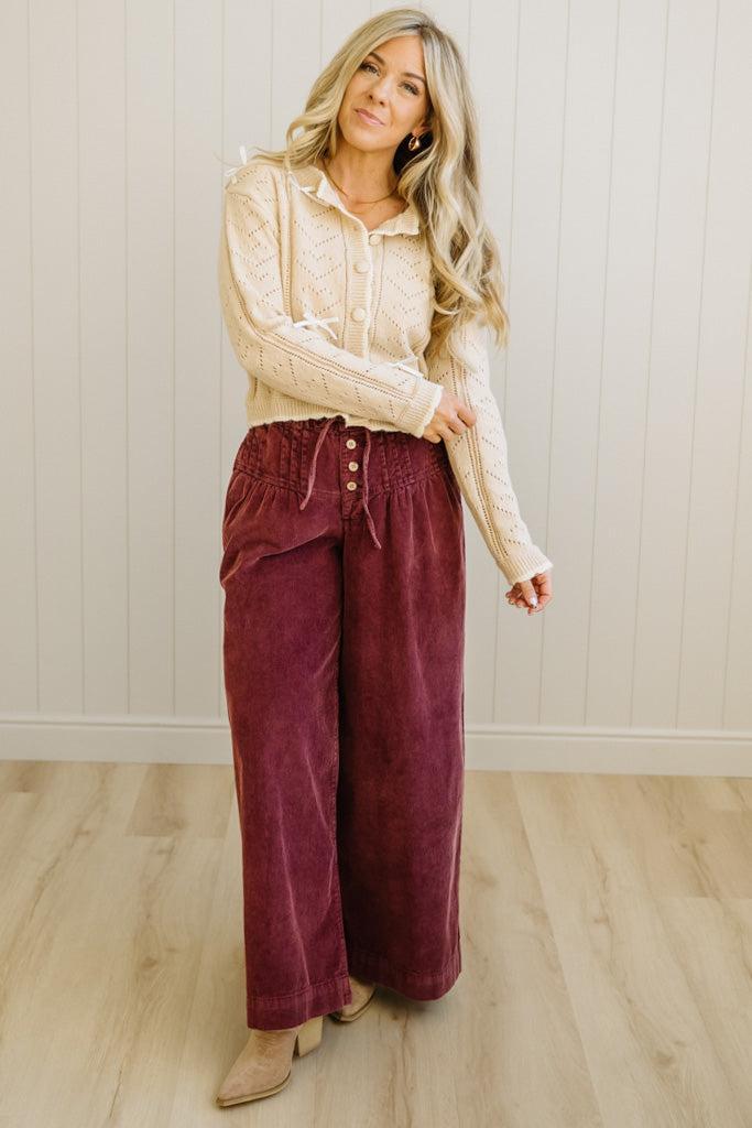 Woman wearing a beige blouse and burgundy pants standing against a white wall.