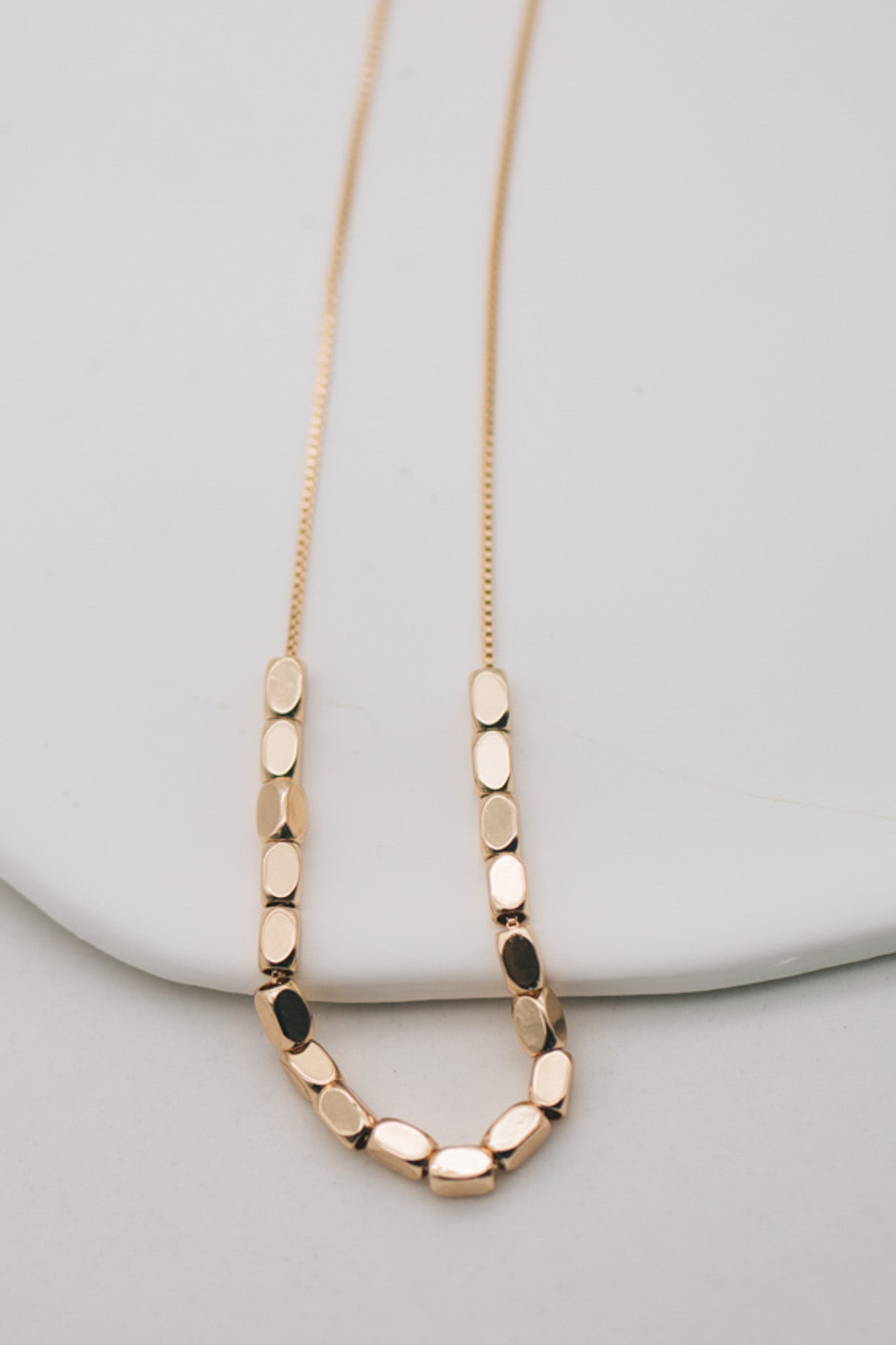 A minimalist beaded necklace with gold plating and small gold beads, displayed on a white plate with a stone background.