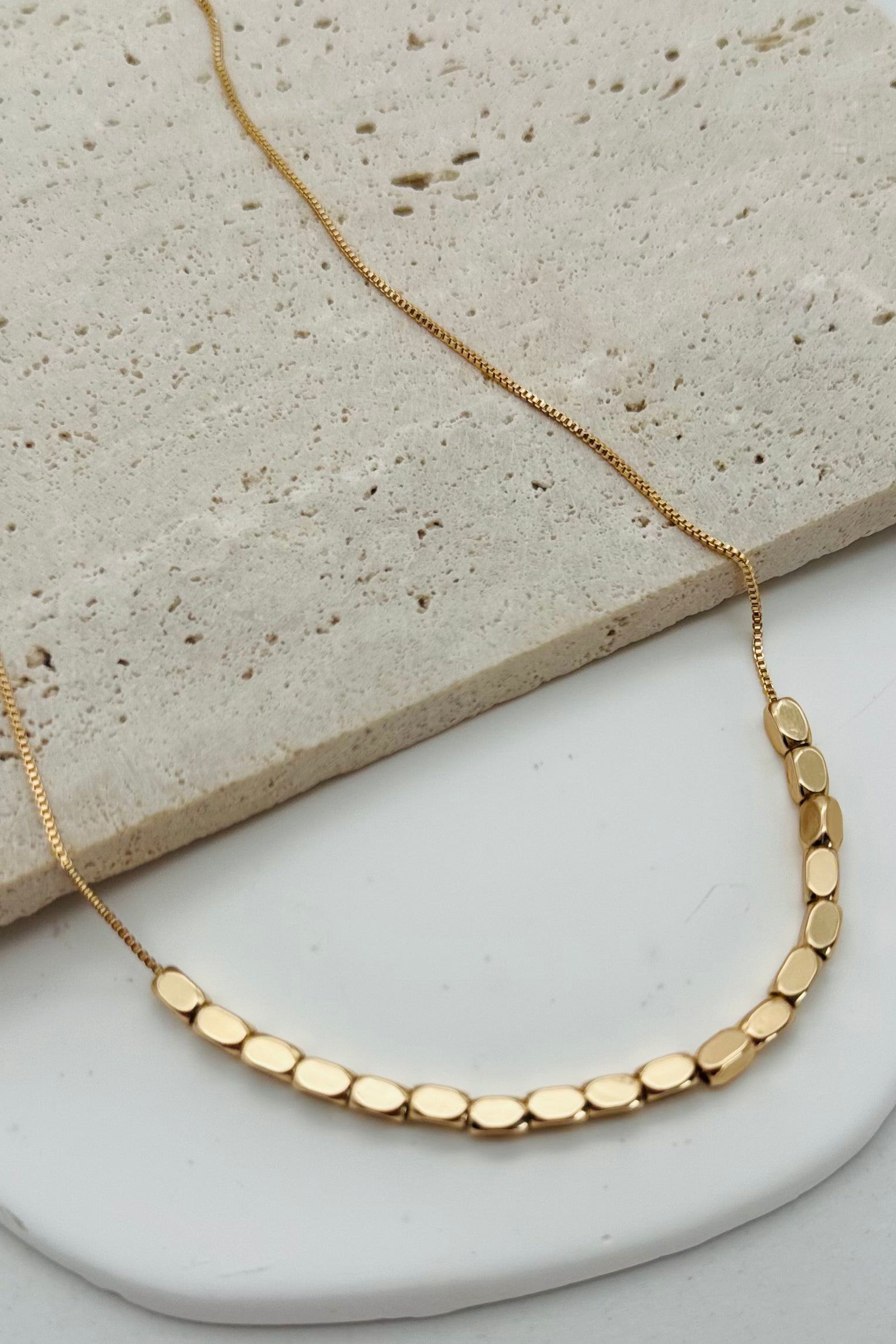 A minimalist beaded necklace with gold plating and small gold beads, displayed on a white plate with a stone background.