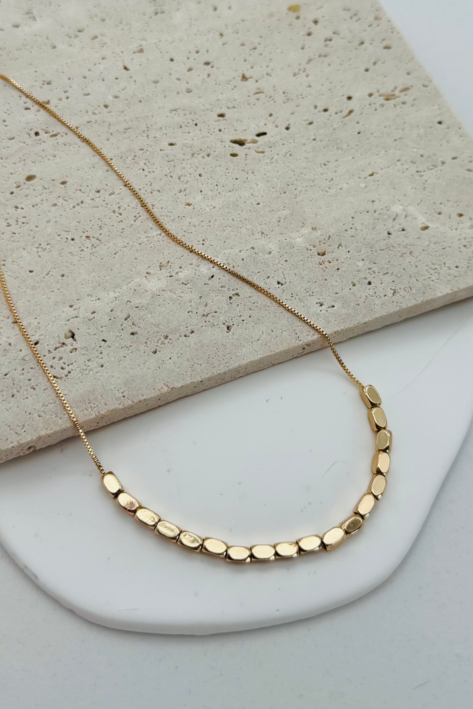 A minimalist beaded necklace with gold plating and small gold beads, displayed on a white plate with a stone background.