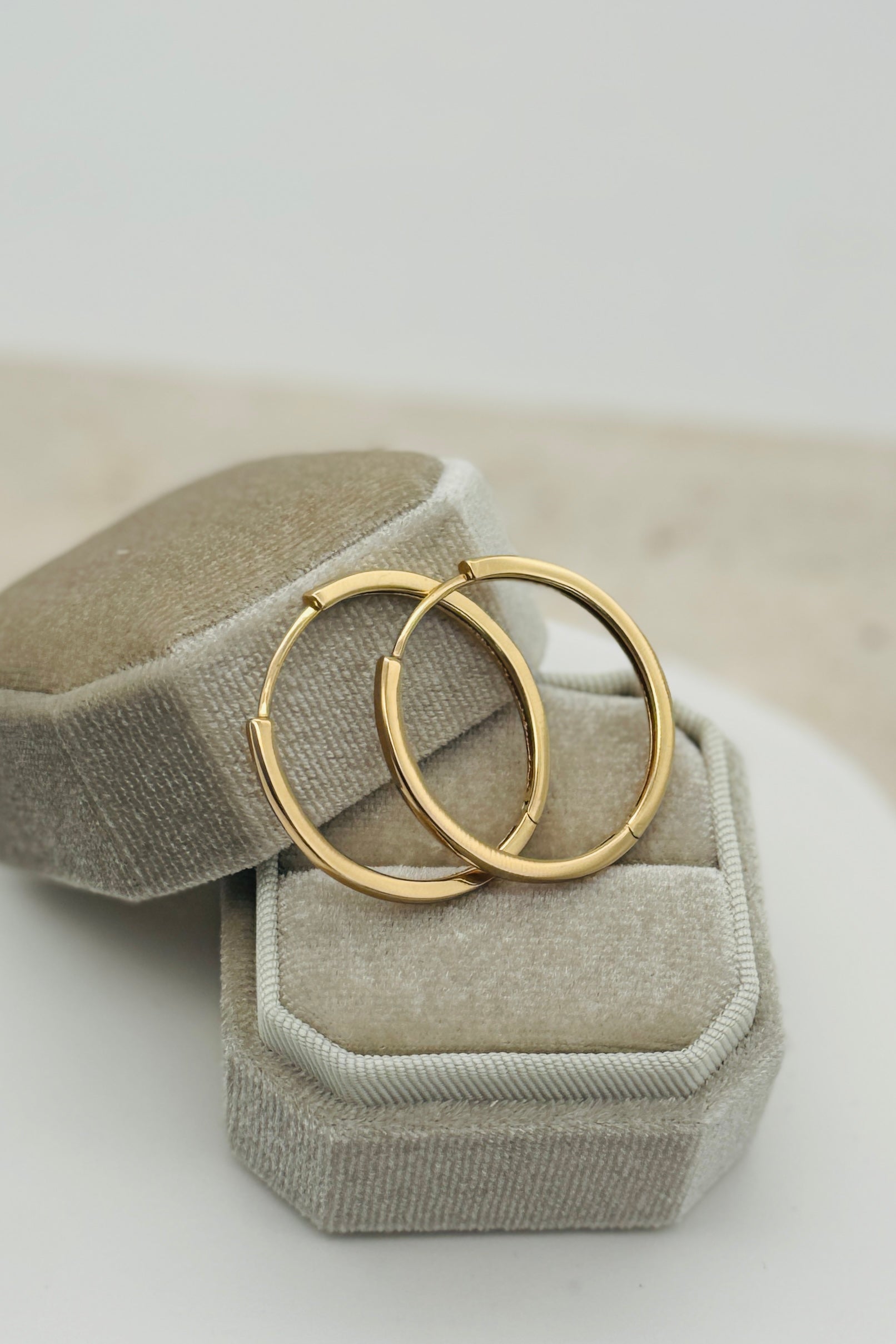 A pair of gold-colored minimalist hoop earrings with a square edge design, displayed on a gray cushion.