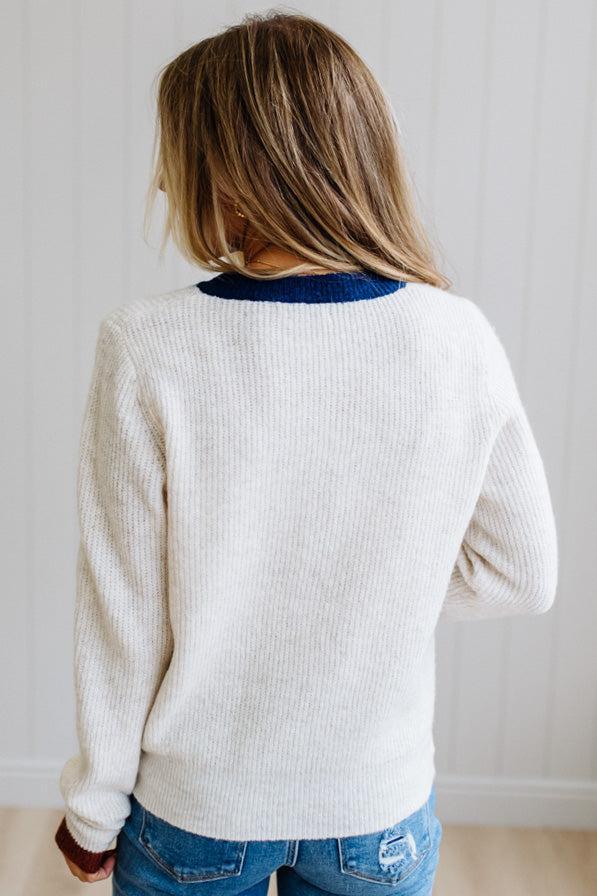 Blonde woman modeling an oat sweater with navy collar, mocha cuffs, and a mocha henlwy with multi color sweater covered buttons. Standing on a light wood floor with light backdrop.