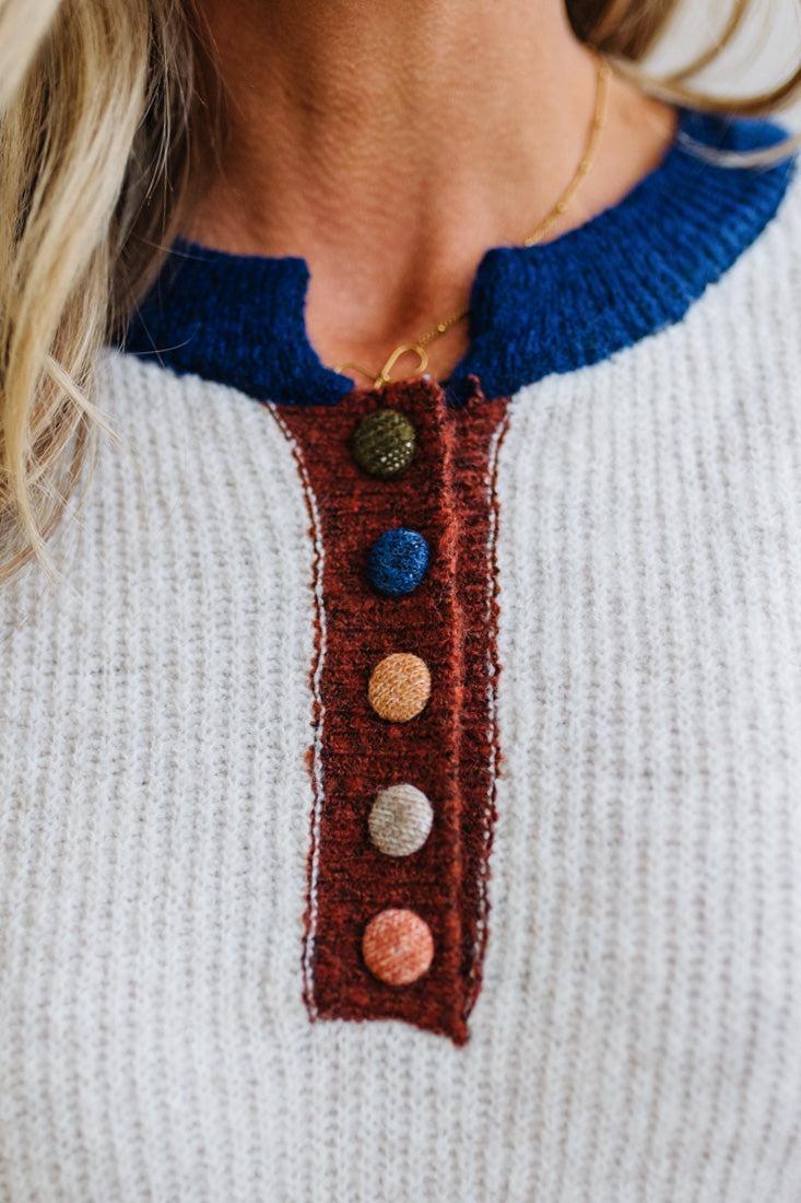 Blonde woman modeling an oat sweater with navy collar, mocha cuffs, and a mocha henlwy with multi color sweater covered buttons. Standing on a light wood floor with light backdrop.