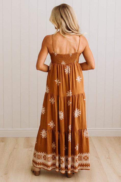 A woman viewed from behind wearing a rust-brown tiered maxi dress with white floral embroidery and a patterned hem, standing against a white paneled wall.