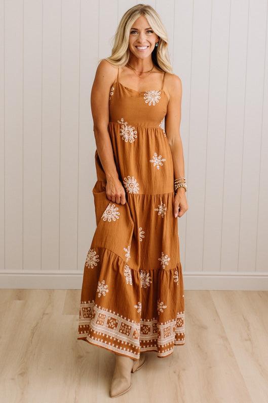 A woman modeling a rust-orange tiered maxi dress with white floral and geometric embroidery, standing against a white paneled wall and light wood floor.