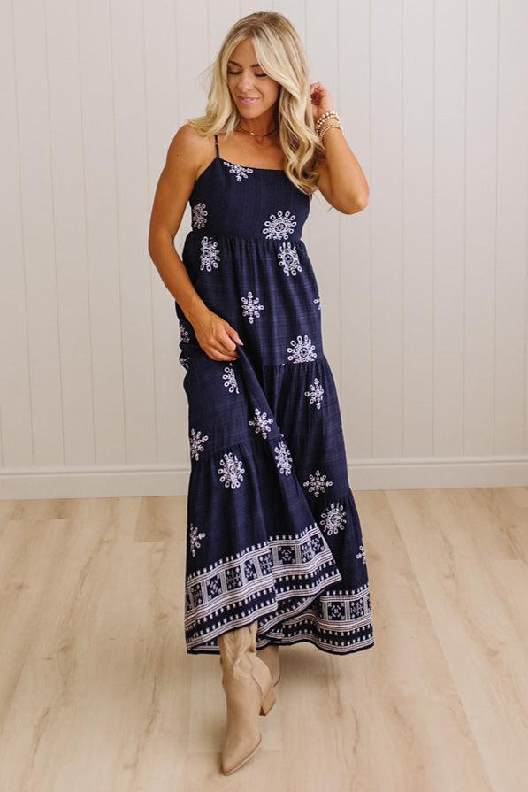 A woman modeling a navy blue maxi dress with white floral and geometric patterns, paired with beige ankle boots, standing in a room with light wood floors and white paneled walls.