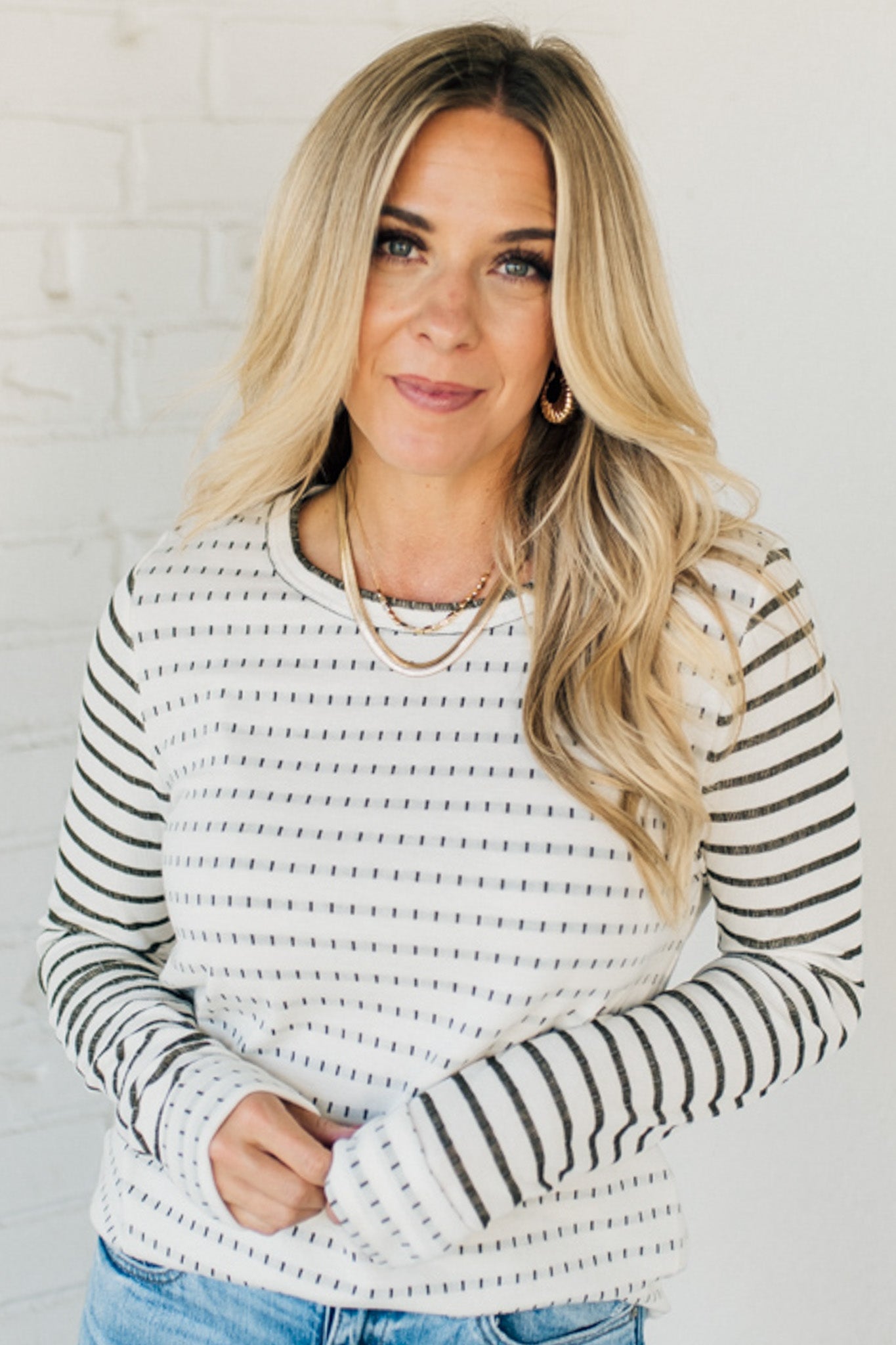 Woman wearing a striped sweater and blue jeans against a white brick wall.