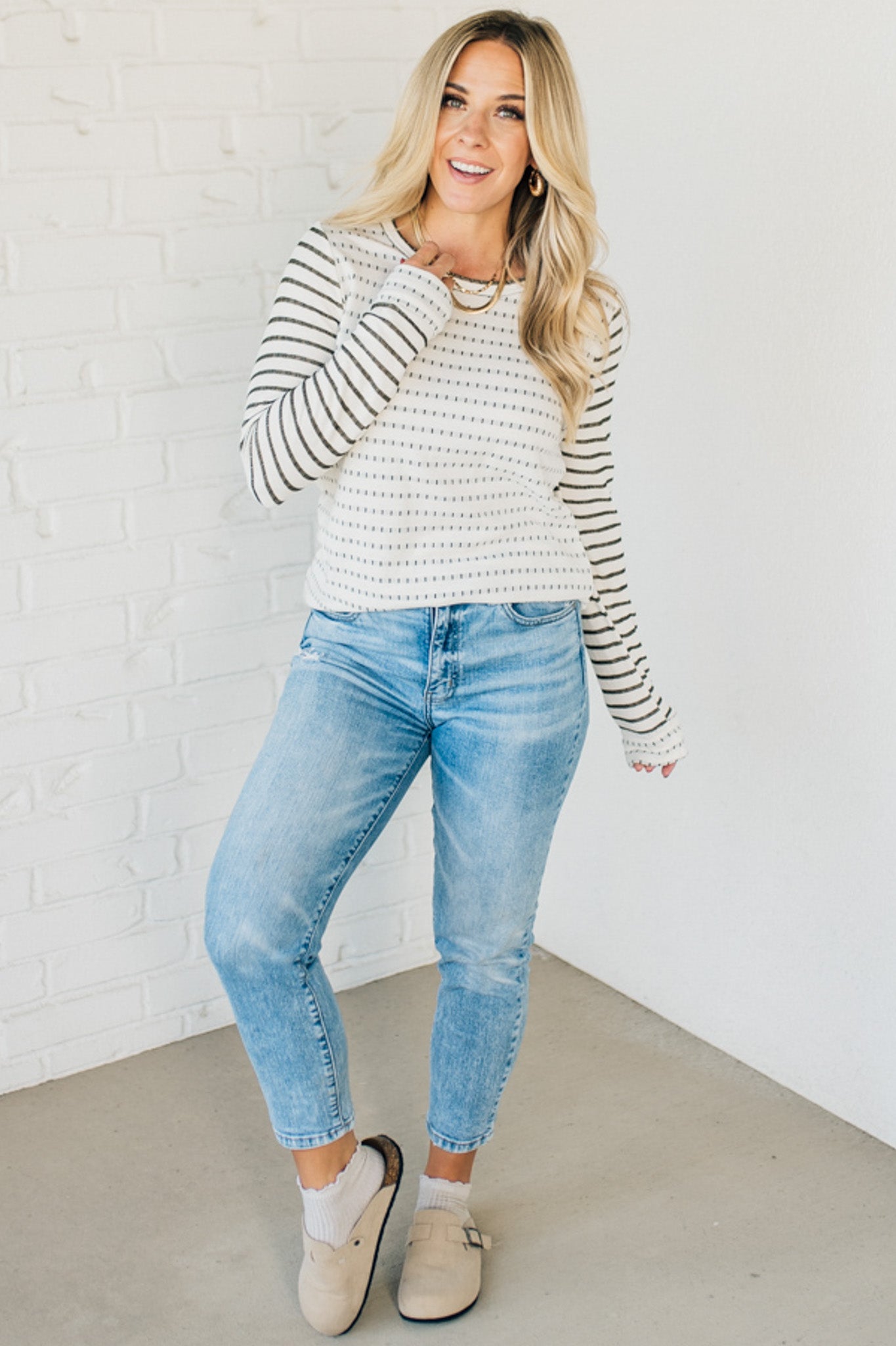 Woman wearing a striped sweater and blue jeans against a white brick wall.
