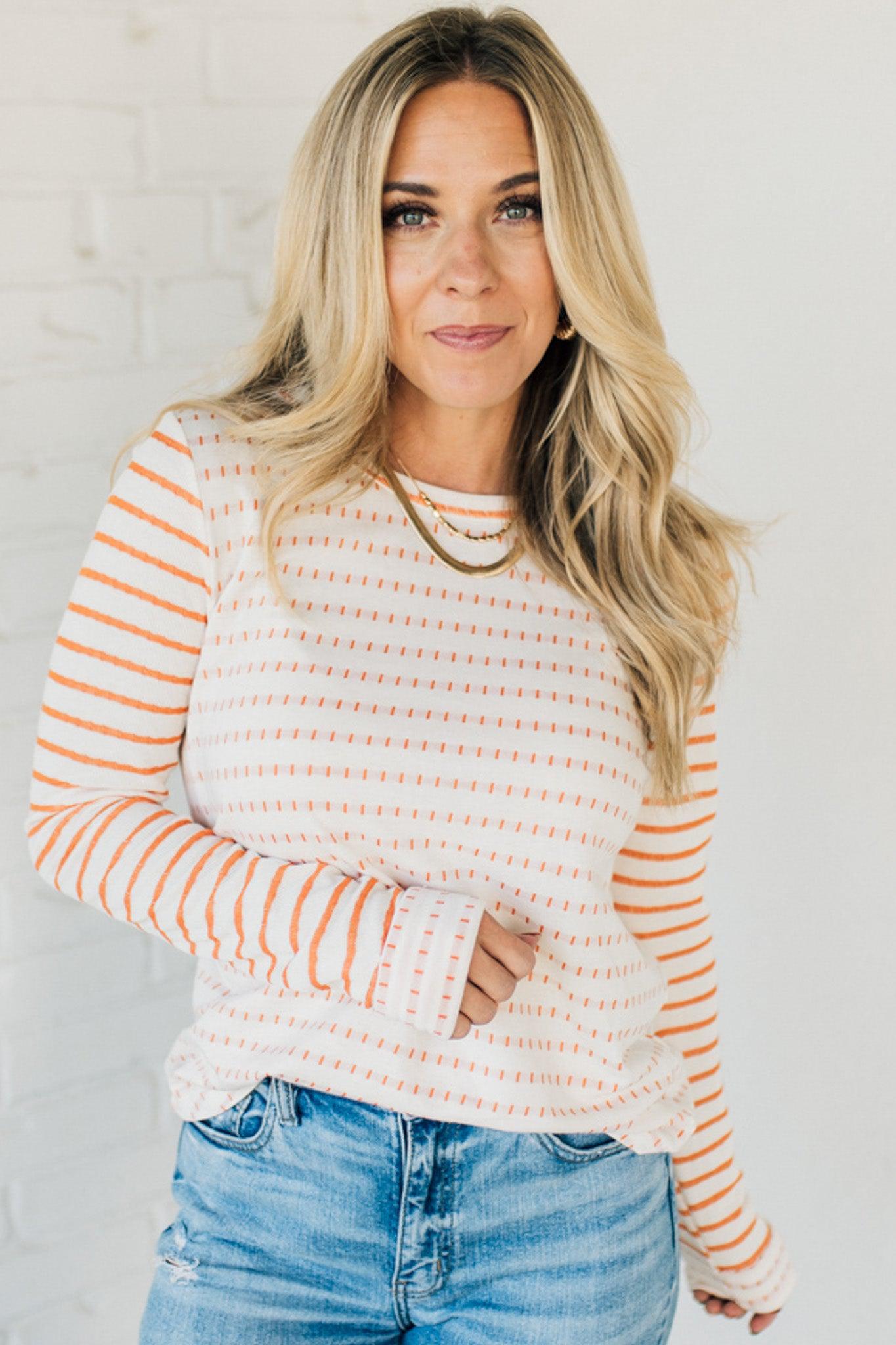 Woman wearing a striped sweater and blue jeans against a white brick wall.