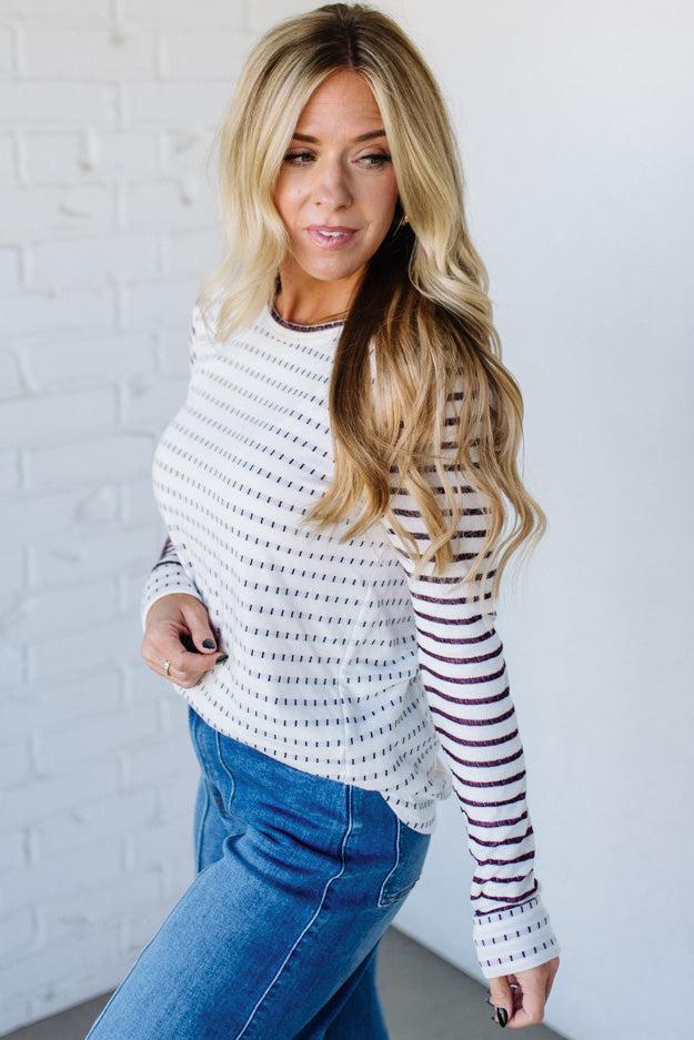 Woman wearing a striped sweater and blue jeans against a white brick wall.