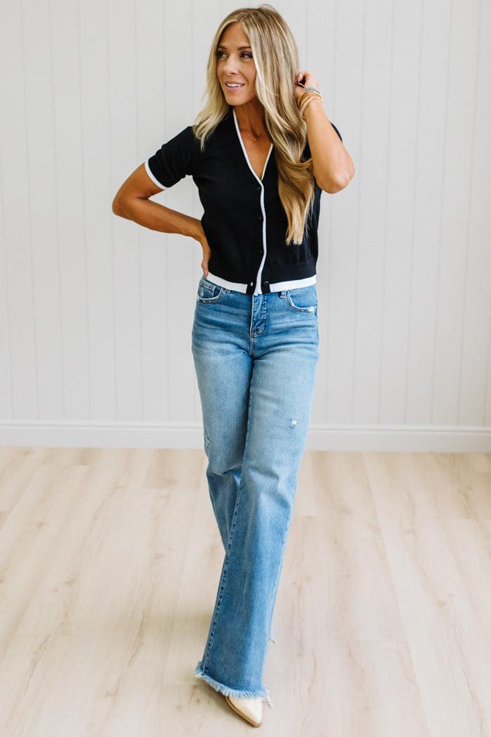 A woman modeling a black short-sleeve knit top with white trim and a white zipper, paired with light-wash blue jeans, standing in a bright room with white paneled walls and light wood floors.