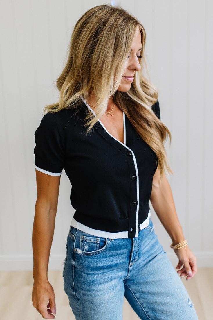 A woman wearing a black short-sleeve button-front top with white trim and light blue jeans, standing against a light indoor wall.