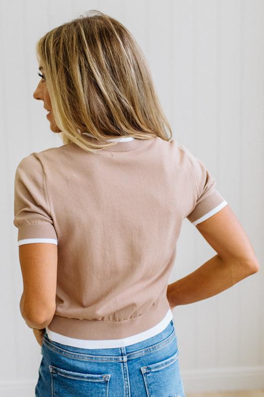 A woman shown from behind wearing a light brown ringer t-shirt with white trim on the collar and sleeve cuffs, paired with blue jeans.