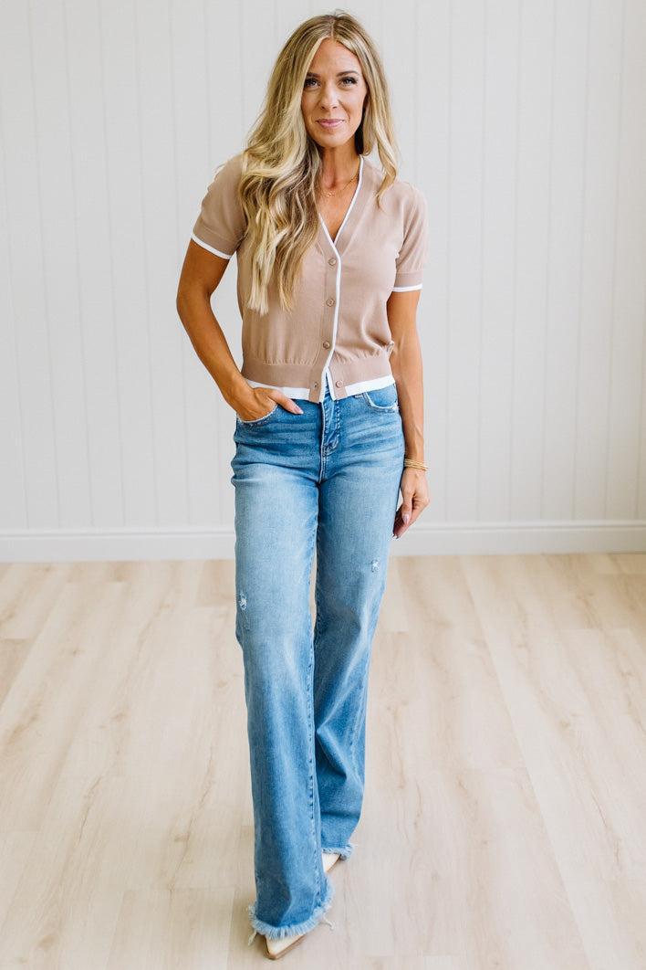 A woman modeling a beige short-sleeve button-front cardigan with white trim and light-wash blue jeans, standing in a bright room with white paneled walls and light wood flooring.