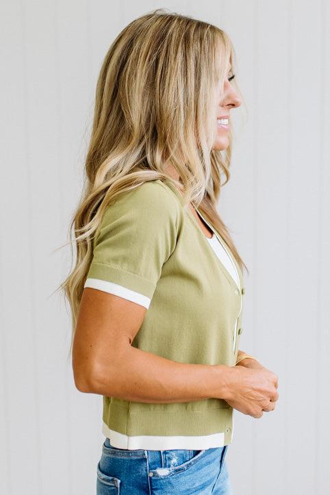 A woman in side profile wearing a sage green short-sleeve knit top with white contrast trim on the sleeves and hem, paired with blue jeans, against a light gray paneled wall.