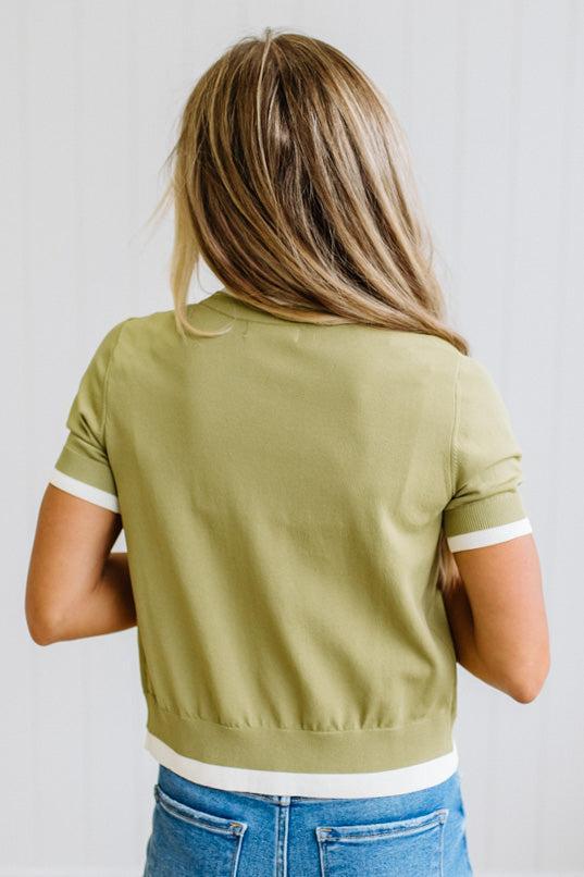 A woman viewed from behind wearing a cropped sage green short-sleeve top with white ribbed trim at the collar, cuffs, and hem, paired with blue jeans against a white paneled wall.