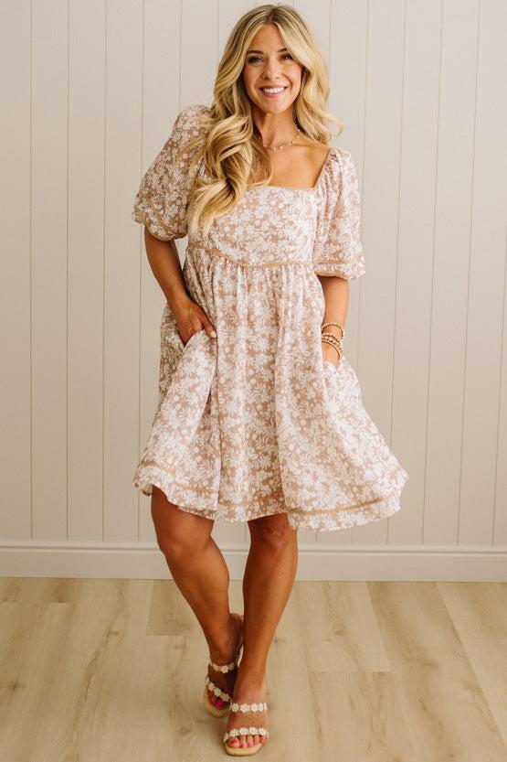 Woman wearing a floral dress standing against a light wooden paneled wall.