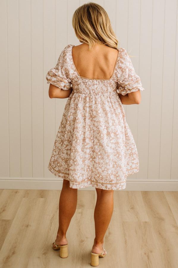 Woman wearing a floral dress with a backless design, standing in a room with wooden flooring and a light-colored wall.