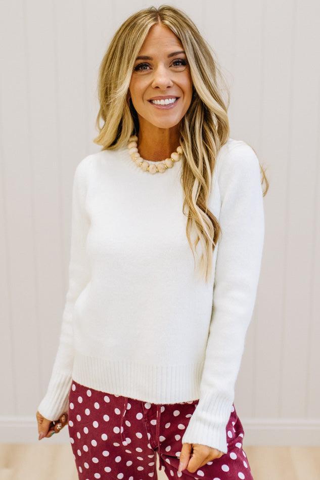 A smiling blonde woman wearing a white long-sleeve knit sweater and burgundy pants with white polka dots, standing against a light paneled wall.