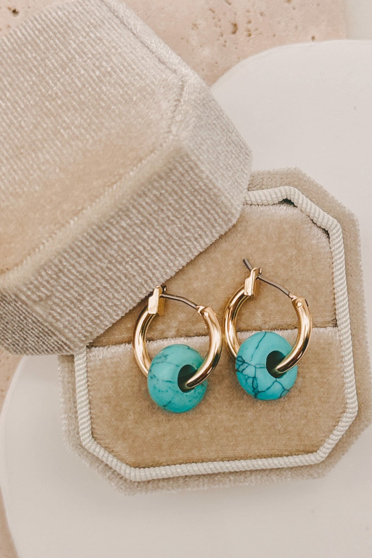 Gold hoop earrings with round turquoise stone pendants, displayed in a beige jewelry box.