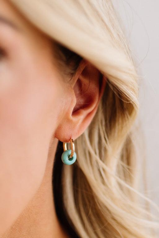 A close-up side view of a woman's ear wearing a small gold hoop earring with a dangling mint green ring charm.