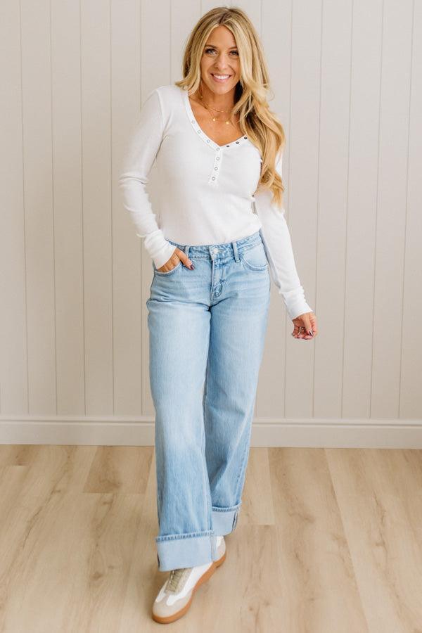 Woman wearing a white long-sleeve top and light blue jeans standing against a beige wall.