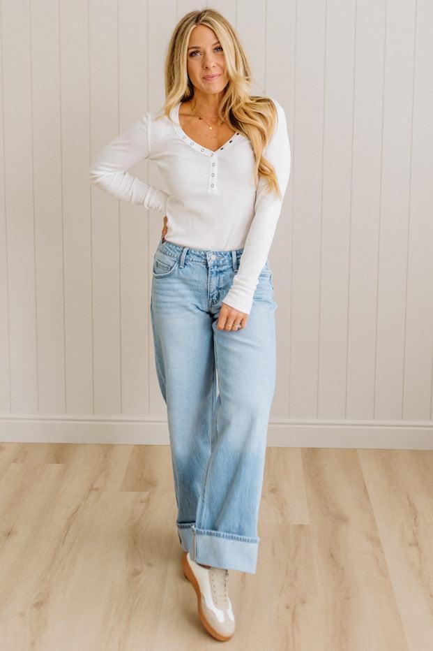 Woman wearing a white blouse and light blue jeans standing against a plain wall.