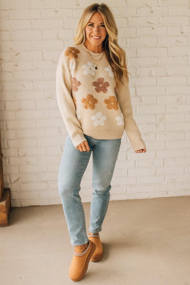 Woman wearing a floral sweater and jeans standing against a white brick wall.