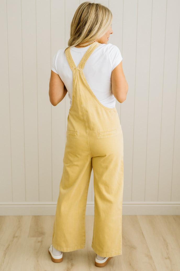 Woman wearing yellow overalls and a white shirt against a light wooden paneled wall.
