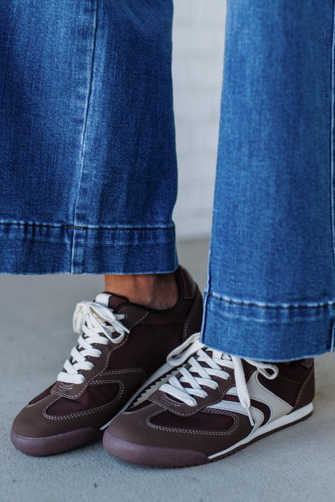 Brown sneakers with white laces worn with blue jeans on a neutral background