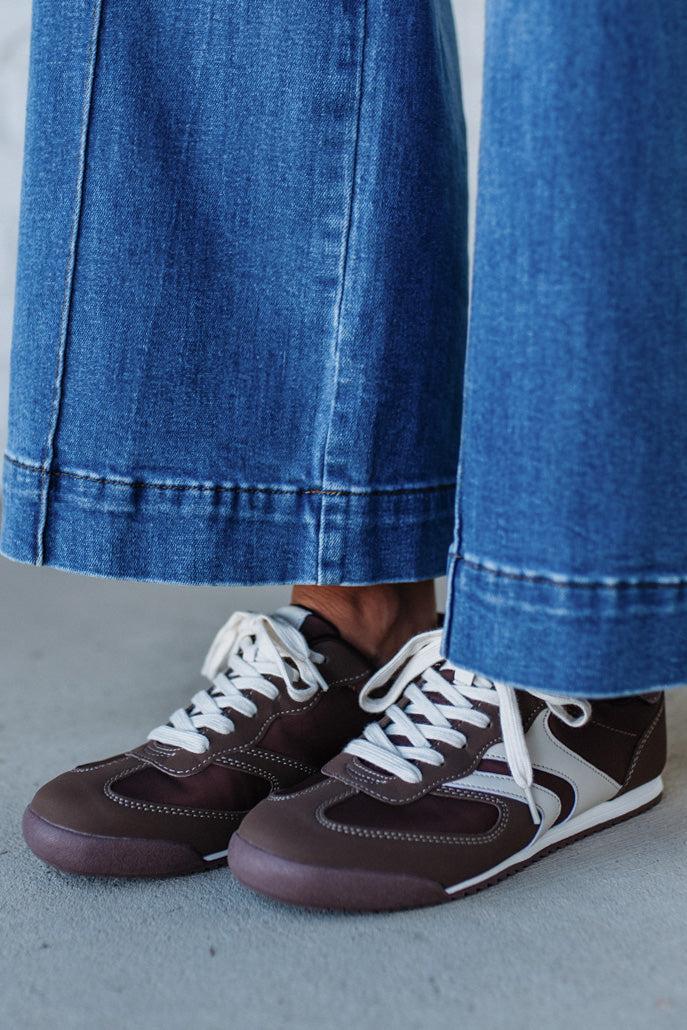 Brown sneakers with white laces worn with blue jeans on a concrete floor.