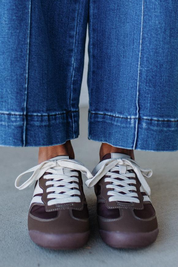 Brown sneakers with white laces worn with blue jeans on a neutral background
