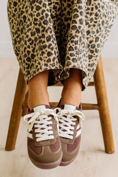 Brown sneakers with white laces worn with leopard jeans on a light wood floor.