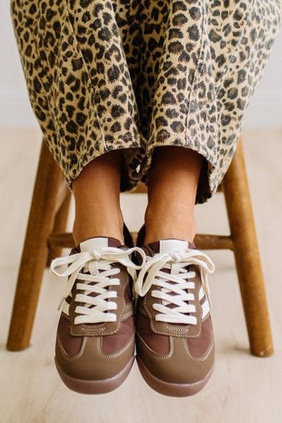 Brown sneakers with white laces worn with leopard jeans on a light wood floor.