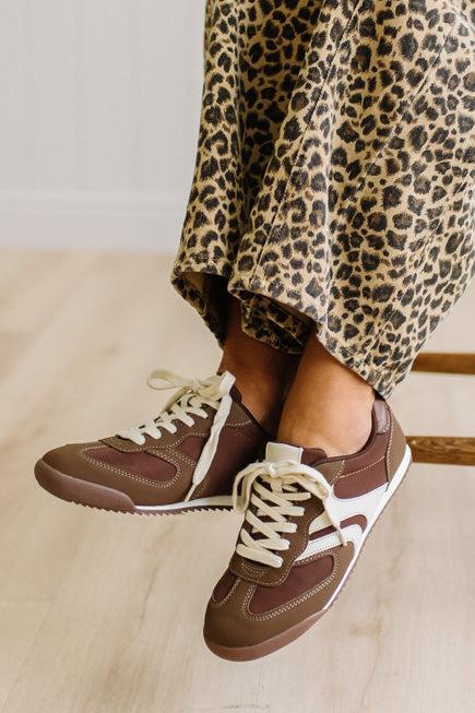 Brown sneakers with white laces worn with leopard jeans on a light wood floor.