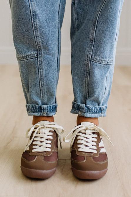 Brown sneakers with white laces worn with leopard jeans on a light wood floor.