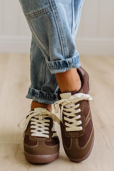 Brown sneakers with white laces worn with leopard jeans on a light wood floor.