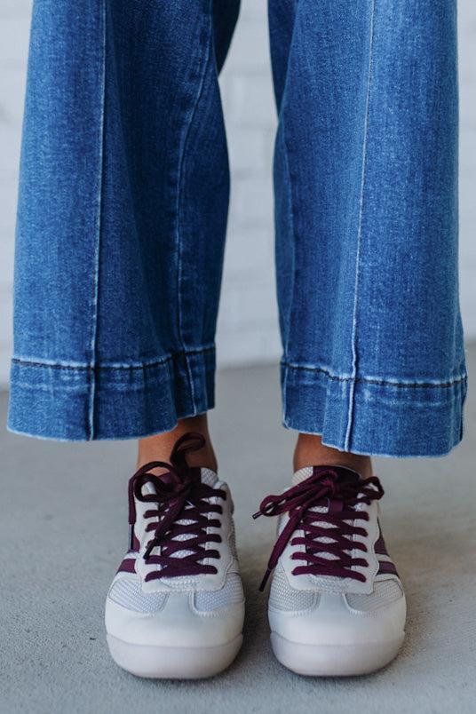 White sneakers with maroon laces worn with blue jeans on a neutral background