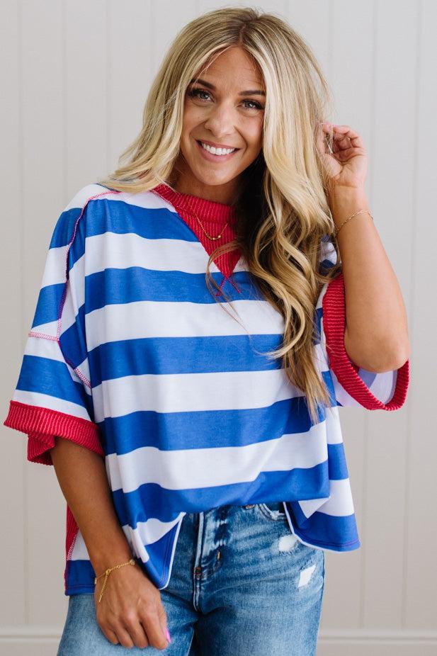 Woman wearing a blue and white striped shirt with red accents, smiling against a light wooden panel background.