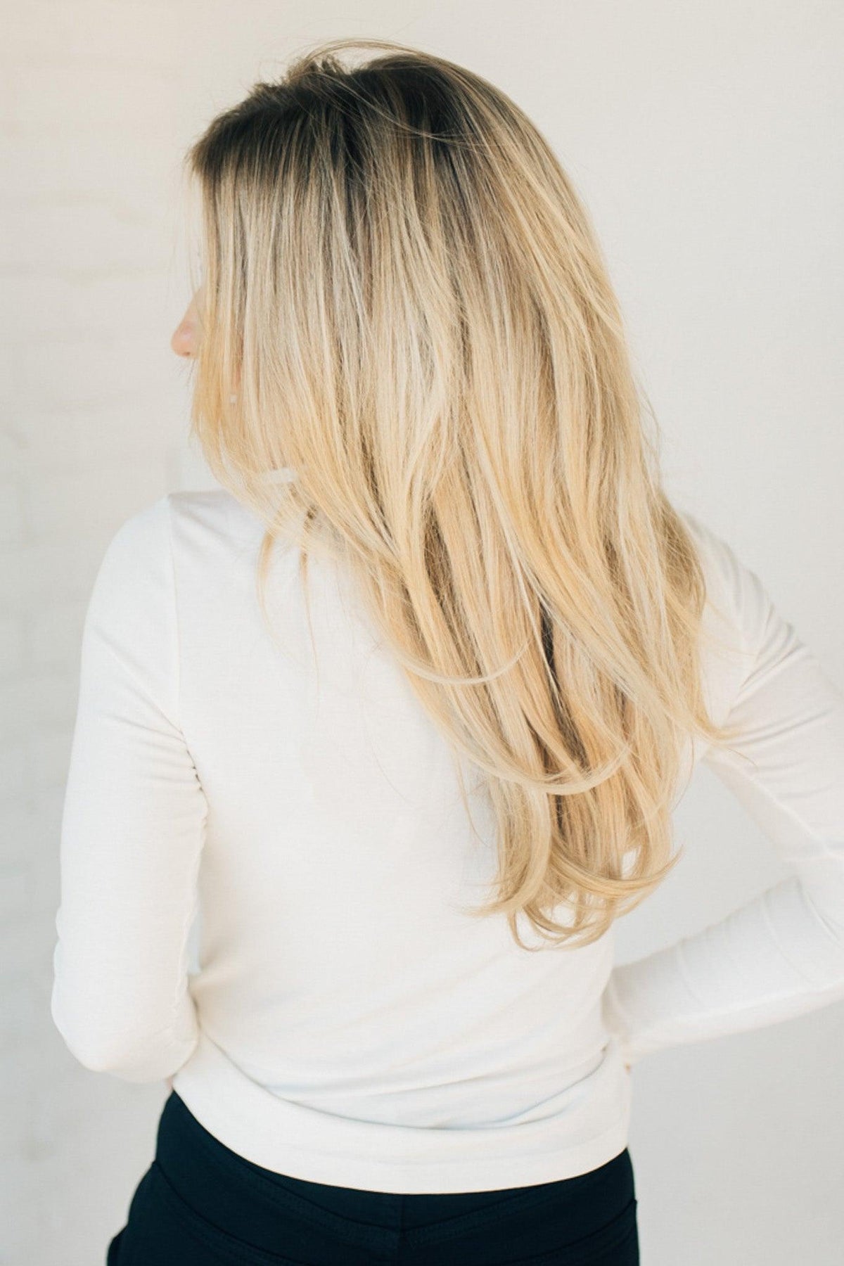 A smiling blonde woman wearing a long-sleeve crewneck knit top in a white color, paired with black jeans, standing against a light neutral paneled wall.